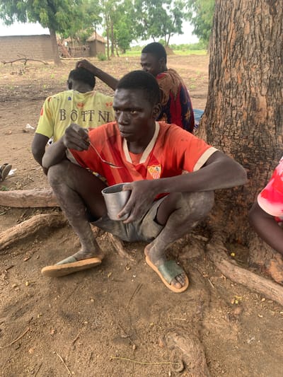 In Dull A, Nigeria, a group of individuals is gathered outdoors, near a large tree with visible roots. The ground is dirt and scattered with dry leaves and twigs. In the foreground, a person wearing an orange and white striped shirt and dark shorts is seated on the ground, facing the viewer. They are holding a metal bowl in their left hand and a spoon in their right, appearing to be eating. Their legs are dirty, and they are wearing a light-colored sandal on their left foot. Behind them, another person in a yellow shirt with black lettering is seated, with their back to the camera, and their head being touched by a hand from another person in the background. A third person, partially visible in the background, is wearing a patterned, colorful fabric. Further behind, traditional mud-brick buildings with thatched roofs are visible amidst trees.