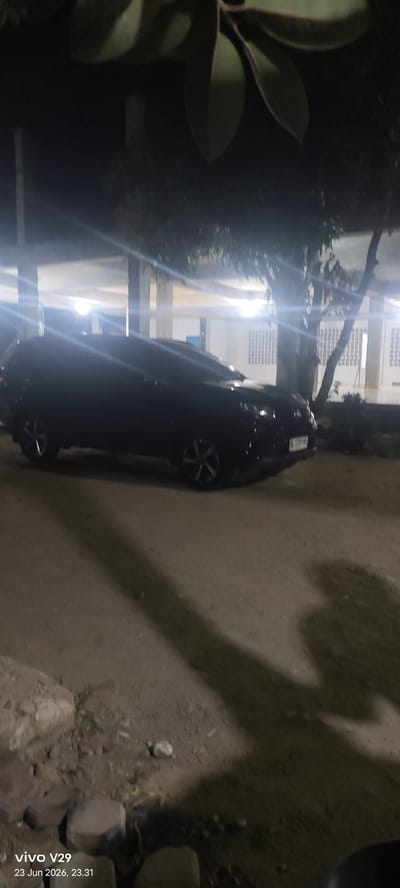 The image depicts a dark-colored SUV parked outdoors at night. The vehicle is the primary subject, positioned centrally in the frame. The setting appears to be a paved or dirt lot adjacent to a building with visible windows and overhead lighting. Trees are present in the background, with leaves from a plant or tree obscuring the top portion of the image. The lighting is artificial, suggesting it's nighttime, and the bright light sources create lens flares. The ground is rough and uneven, with some rocks and debris visible. A long shadow stretches across the ground from the vehicle and possibly other unseen objects.
The timestamp at the bottom left corner indicates the image was taken on "23 Jun 2026, 23.31" by a "vivo V29" device. The location context provided is Meunasahaluedrienlhoksukon Barat, Indonesia. No people are visible in the image. The overall mood is dim and somewhat underexposed due to the nighttime setting and the camera's exposure choices. The activity is simply parking, with no discernable action or event taking place beyond that. The visible text confirms the date, time, and device used for the photograph.