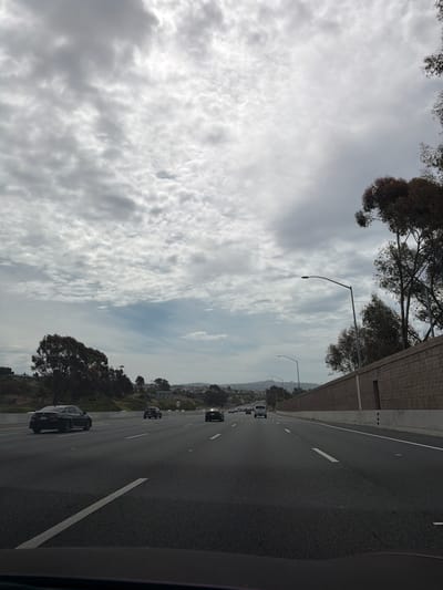 The image captures a view from a vehicle traveling on a multi-lane highway. The sky is overcast with dense, textured clouds. On the right side of the highway, a textured brick retaining wall runs alongside the road, with trees and vegetation visible beyond it. On the left side, there are rolling hills with scattered trees and buildings in the distance. Several cars are visible on the highway, all moving in the same direction. The road itself is asphalt with clear white lane markings. Streetlights are present along the highway. The scene suggests a cloudy day in an urban or suburban area, possibly San Clemente, California, given the typical coastal highway architecture and vegetation.