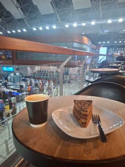 The image shows a slice of chocolate cake and a coffee cup on a wooden table. In the background, there is a view of an airport security checkpoint area in Pendik, Turkey, with people going through security. The overall setting appears to be a cafe or seating area overlooking the airport operations. The lighting is bright, typical of an airport environment.