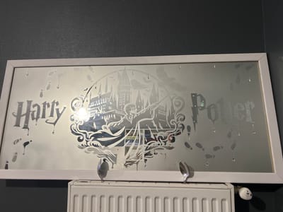 A rectangular mirror with a white frame hangs on a grey, textured wall. The mirror's surface is etched with a design featuring the words "Harry Potter" in a dripping font, the Hogwarts castle, a silhouette of Harry Potter casting a spell, and footprints. There are small, clear droplets of liquid scattered across the mirror's surface. Two spoons are placed on the top edge of a white radiator situated below the mirror. The radiator has a white thermostatic valve on its right side. The scene is located in Rozérieulles, France.