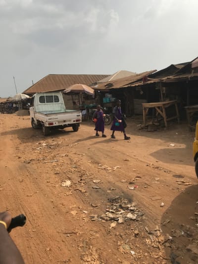 The image depicts a bustling street scene in Ogbese, Nigeria, under an overcast sky. A wide, unpaved dirt road, littered with debris and tire tracks, dominates the foreground and mid-ground.

In the mid-ground, a white compact pickup truck with a light blue bed liner is parked facing away from the viewer on the left side of the road. Two young females, likely schoolgirls, wearing matching purple uniforms with black shoes and socks, walk from left to right across the road in front of the truck. The girl on the left carries an orange item, and the girl on the right carries a white or light-colored container or bag. They cast discernible shadows to their left.

To the right of the walking girls, the edge of a yellow vehicle is visible. Along both sides of the road, particularly in the background, numerous market stalls and single-story structures with corrugated metal roofs are present. Several stalls feature large shade umbrellas, one tan and one white. Visible items in the market include baskets, crates, and wooden tables used for display. Further down the road on the left, other individuals and motorcycles are present near additional market activity. A bare arm with a yellow band is partially visible in the bottom left corner of the frame.