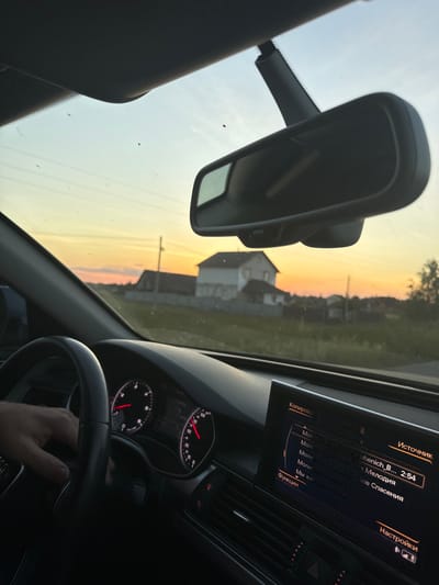 This media file shows the interior of a car, viewed from the driver's perspective, during what appears to be sunset. The dashboard is visible, including a car's infotainment system displaying Russian text that translates to "Source," "Lubenich_B... 2:54," "Molotov," "Molot Melody," "Molitva - Call of Salvation," "We live," "Functions," and "Settings." The infotainment system also shows a list of songs, with the current track being "Lubenich_B..." which is 2 minutes and 54 seconds long.
The driver's hand is on the steering wheel, suggesting they are driving. The windshield shows a rural landscape with houses and trees, bathed in the warm, golden light of sunset, indicating it is either early morning or late evening. The sky displays a gradient of yellow, orange, and pink hues, typical of a sunset. The overall scene is somewhat blurry, possibly due to the movement of the car or low light conditions. The location is identified as Dovhe, Ukraine.