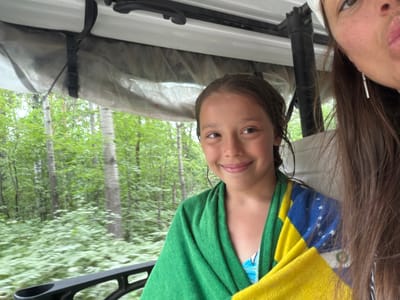 This media file shows a selfie taken from inside a golf cart as it drives through a forest. The primary subject is a young girl in the foreground, wrapped in a green towel and a towel with a yellow, blue, and green pattern. She is looking towards the camera with a smile. To her right, partially visible, is an adult woman, also looking at the camera with her lips pursed. The background is a blur of green trees and foliage, suggesting the cart is in motion. The presence of the towels and the setting imply they may have just been swimming or are on their way to or from a body of water. The lighting suggests it is daytime, possibly a cloudy or overcast day given the soft light. The scene appears to be a natural, wooded area, likely a recreational spot. There is no visible text in the image.