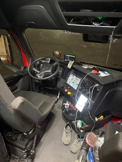 The image shows the interior of a truck cab, likely in New Britain, United States of America, as indicated by the provided context. The viewpoint is from the passenger side, looking towards the driver's side and the dashboard.
The driver's seat is a dark grey fabric material with visible stitching and an armrest. The seatbelt is fastened with a red buckle. The floor of the cab is dark grey, with a textured surface. There are two pairs of grey athletic shoes on the floor, one pair near the driver's seat and another pair further back.
The dashboard is black and features multiple electronic devices. Two GPS units are mounted on the dashboard, one smaller unit at the top and a larger tablet-like device below it, displaying a map. Various cables and chargers are connected to these devices. A red object, possibly a portable charger, is visible on the dashboard. A digital display screen is also visible on the lower part of the dashboard. Several buttons and controls are present on the steering wheel and the center console.
The steering wheel is black and has integrated controls. The interior of the truck appears somewhat cluttered, with personal items visible on the dashboard and floor. A red interior door panel is visible on the left side of the image, and a side mirror is also visible through the window. Above the dashboard is a storage compartment, containing various items including what appears to be a wallet and electronic accessories.
The lighting suggests it might be nighttime or a dimly lit indoor environment. The background is dark, with some indistinct illuminated areas, possibly indicating a parking garage or an industrial setting.