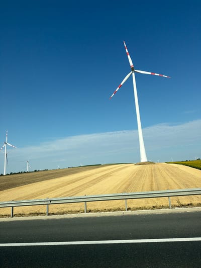 The image depicts a wind farm situated in the agricultural landscape near Wilfersdorf, Austria. In the foreground, a section of an asphalt road with a white line is visible, bordered by a silver metal guardrail. Beyond the guardrail lies a harvested field of golden-yellow stubble, meticulously patterned with parallel rows. In the mid-ground, several white wind turbines with red and white striped blades stand tall against a clear, vibrant blue sky. One prominent turbine is positioned on the right side of the frame, its tower casting a shadow on the field. Further in the background, more turbines are scattered across the horizon, interspersed with patches of green vegetation and what appears to be a field of sunflowers on the far right. The scene is bathed in bright daylight, suggesting a sunny day.