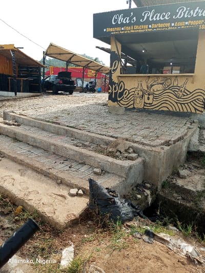 The image shows a set of concrete stairs leading down to a drainage ditch in Ado Ekiti, Nigeria, located at coordinates 7.649 latitude and 5.226 longitude. The stairs are partially broken and overgrown with weeds. A black plastic bag sits at the base of the steps near the ditch. The steps are made of concrete blocks laid in a pattern. The steps are surrounded by a dirt area with some green grass growing. In the background, a restaurant with a sign that reads "Oha's Place Bistro" is visible. The restaurant is located on a paved area. A person is visible inside the restaurant. The image appears to be taken at a low angle, looking up at the steps. The sky is overcast and gray. The image appears to be taken in the late afternoon or early evening. There is a small amount of litter in the image.