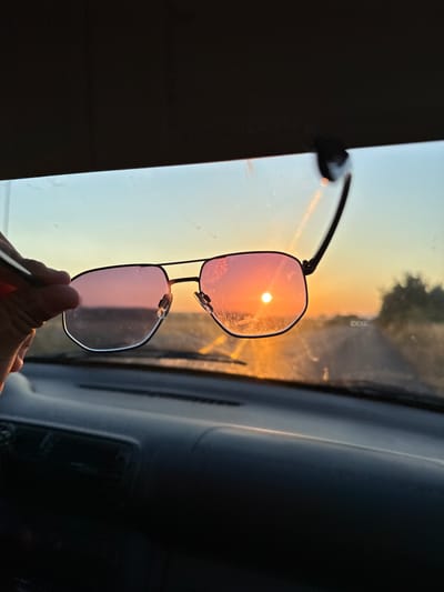 A person's hand holds a pair of glasses up to a car's windshield during sunset. The sun, a bright orb, is visible through the upper lens of the glasses, casting a warm, pinkish glow on the lens and the surrounding scene. The background shows a blurred landscape with trees and a road, illuminated by the setting sun. The interior of the car, including the dashboard and window frame, is visible on the right side of the image, rendered in dark tones. The overall impression is one of a peaceful evening commute or a moment of reflection. The time of day is clearly sunset.