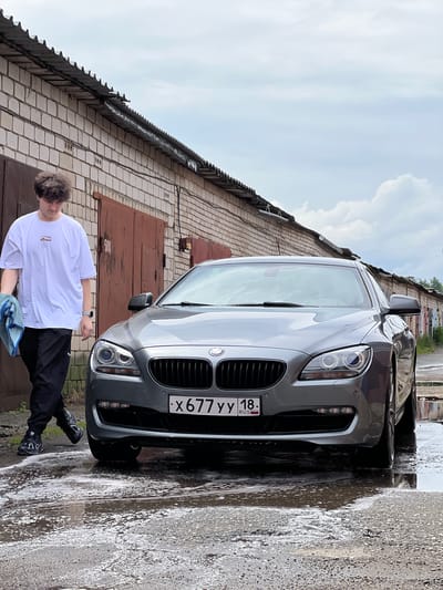 A young man in a white t-shirt and black pants is washing a gray BMW car parked on a wet, gravelly surface. The man is holding a blue microfiber cloth, suggesting he is in the process of cleaning the vehicle. The scene appears to be outdoors, with a brick building and garage doors in the background, indicating a residential or industrial area. The sky is overcast, with visible clouds, suggesting a cloudy day. There is water and soap suds on the ground around the car, confirming the car washing activity. The license plate on the car reads "X677YY 18 RUS." The overall mood seems casual and task-oriented, with the man focused on his chore. The location context provided is Veteran-2, Russia.