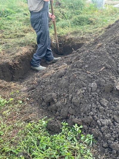 A person stands in a freshly dug trench, wielding a garden fork. The trench runs through soil covered with dry grass and some green weeds. A large pile of dark soil sits next to the trench. The person wears dark blue pants and rubber boots. The location is likely an outdoor garden or field environment. The exact location of Makariopolsko, Bulgaria, cannot be determined from the image.
