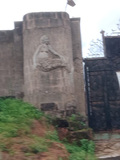 A partially blurred, medium shot captures an outdoor scene of a weathered, concrete structure with a bas-relief carving and a dark metal gate. The carving on the concrete wall depicts a stylized, nude figure in a reclining or seated position, with the details of the face and body indistinct due to the blur. Adjacent to the concrete structure is a tall, ornate, dark metal gate, partially obscuring a lighter colored area behind it. To the left of the concrete structure, a grassy slope with visible red earth beneath the vegetation descends. The sky above is overcast and white. The overall impression is of a slightly aged or neglected entrance to a property in a tropical or sub-tropical environment, given the vegetation. The location is identified as Iju, Nigeria, suggesting a context of local architecture and artistry.