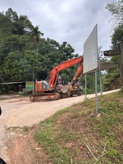 The image depicts an excavator positioned on a dirt and concrete road in Cổ Lũng Commune, Vietnam. The excavator, a large orange machine with a metallic bucket, is stationary and appears to be in a working or parked state. To the left of the excavator, a small, rustic-looking structure with a corrugated metal roof is visible, suggesting a rural or agricultural setting. A green sign with text and illustrations stands near the excavator, indicating information or instructions. A tall, white sign with some text is positioned to the right of the excavator, further down the road. The environment is characterized by lush green trees and vegetation covering hillsides in the background, under an overcast sky. The ground consists of a mixture of soil and a paved or concrete surface. There are no people clearly visible in the image. The scene conveys a sense of construction or ongoing work in a natural, mountainous landscape.