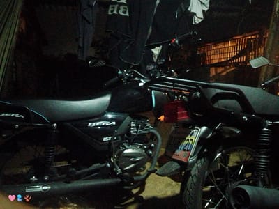 The image shows two motorcycles parked side-by-side in an outdoor setting, possibly in El Cercado, Venezuela. The motorcycles appear to be of the "Bera" brand, with the "150" model number visible on one. The motorcycles are black in color. Clothes are hanging above them on a clothesline. In the background, there is a structure with a barred window. The lighting is dim, suggesting it is night. There is no evidence of other people or activities beyond the presence of the motorcycles and clothesline.