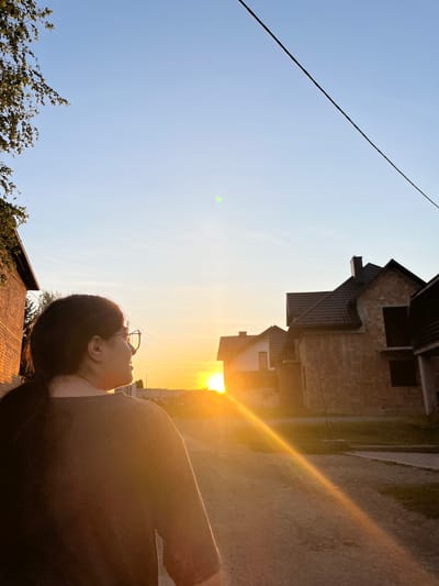 In the city of Koroviia, Ukraine, a person wearing glasses and a dark t-shirt is facing away from the camera, looking towards a sunset. The sun casts a warm, golden light, creating lens flares that streak across the image. The scene appears to be a residential area with houses under construction visible in the background, characterized by brick exteriors and dark roofs. A dirt road or pathway runs through the scene. Overhead, a power line is visible against the clear, pale blue sky. To the left, the branches of a tree with green leaves are partially in frame.