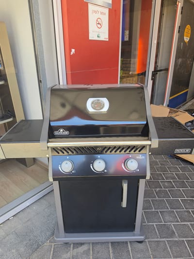 A black gas grill, prominently branded "Napoleon" and featuring the model name "ROGUE," is positioned outdoors on dark gray interlocking paver stones. The grill has a sleek black finish, a temperature gauge on its lid, and three control knobs on its front panel, indicating settings like ignition and flame intensity. One knob features a lightning bolt symbol.
The setting appears to be outside a storefront in Rehovot, Israel, under bright daylight conditions, suggested by the clear reflections and sharp lighting. To the left of the grill is a red wall or door, against which a white sign with a red circle and a crossed-out cigarette is affixed, clearly indicating "אסור לעשן" (No Smoking) in Hebrew. Reflections in the adjacent windows show parts of the building's interior, including what might be lighting or shelves. In the background, behind the grill, several cardboard boxes are visible, suggesting the grill might be newly delivered or displayed. The overall impression is a commercial outdoor space, possibly for retail display.
