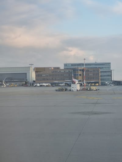The image shows an airport scene in Echterdingen, Germany. The Stuttgart Airport is visible with a building labeled "STUTTGART." An airplane is parked near the terminal, connected to ground support equipment. The weather is overcast. The setting is a concrete apron adjacent to airport buildings.