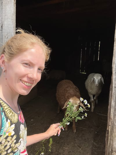 In the city of Сям-Можга, Russia, a young woman with blonde hair and piercings is taking a selfie outside a rustic barn. She is smiling and holding a bouquet of small white daisies. Inside the dark barn, two sheep are visible: a brown one in the foreground and a white one further back. The barn appears to be made of wood, with some sections weathered and aged. The ground inside the barn is dirt, with some scattered hay. The overall scene suggests a rural setting on a sunny day.