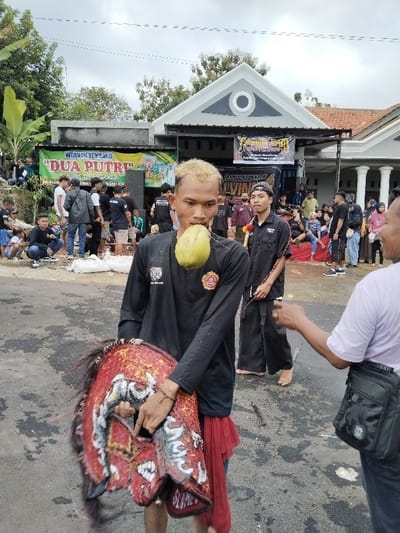 An outdoor scene in Sumberejo, Indonesia, depicts a public gathering. In the foreground, a male with bleached blonde hair, wearing a black long-sleeved shirt with a gold emblem and white text, and a red lower garment, holds a red, white, and black decorated Kuda Lumping horse-head mask in his left arm. A green young coconut is visibly pierced through his cheeks. A dark beaded bracelet is on his left wrist. To his right, a second male, barefoot, wears a black cap, a black collared shirt with a white logo, and black wide-legged pants, holding a thin dark stick. A yellow and red garland is around his neck. A third, partially visible male on the far right, wearing a light shirt and a black fanny pack, extends his right arm towards the central figure. The ground is a dark paved surface with scattered light-colored material. In the midground, a crowd of diverse individuals observes from behind the foreground figures. A green and yellow banner advertising "WARUNG SEMBAKO DUA PUTRI" and a black banner with yellow text are displayed. Audio speakers are visible on the left. The background features a white building with a grey triangular roof and a circular decorative element, adjacent to a grey flat-roofed building and residential structures with tiled roofs. Green foliage, including banana trees, is present under an overcast sky.