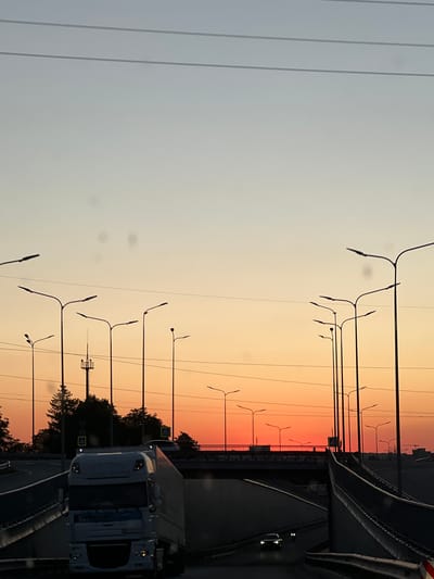 The image depicts a sunset scene on a highway overpass in Velyki Hai, Ukraine. The sky exhibits a gradient of pale blue at the top transitioning to a vibrant orange and red hue near the horizon. Silhouetted against the sky are numerous streetlights and power lines. In the foreground, a white semi-trailer truck is visible on the road, alongside other vehicles. The scene is framed by elevated road infrastructure, including concrete barriers and ramps, suggesting a complex interchange. The overall atmosphere is one of a twilight commute.