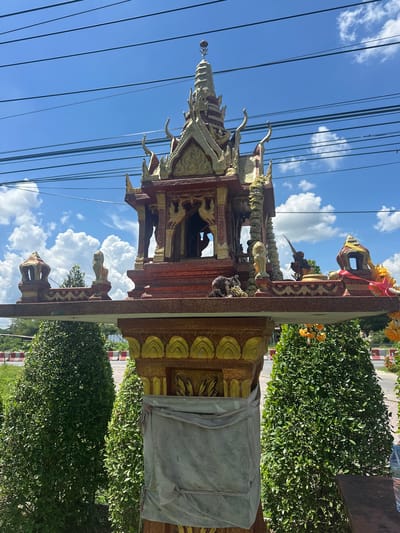 A small, ornate shrine is positioned outdoors in Tambon Phraek Ha, Thailand. The shrine is a multi-tiered structure with red and gold decorative elements, topped with intricate spires. It rests on a pedestal, which is partially draped with a light-colored cloth. Flanking the shrine are two neatly trimmed green bushes. In the background, a bright blue sky with scattered white clouds is visible, crossed by several dark electrical wires. The scene is lit by natural sunlight, indicating it is daytime.