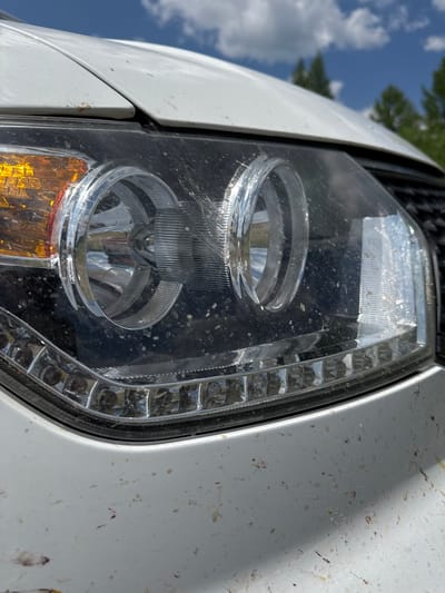This media file is a close-up, low-angle shot of a white vehicle's headlight. The headlight is clear and shows two circular reflectors, one above the other. A row of small, rectangular LED lights is visible along the outer edge of the headlight assembly, and an amber turn signal is situated below the lower reflector. The headlight lens is dusty and smudged with dirt and what appear to be insect remains.
The vehicle's white bodywork, also speckled with dirt, is visible to the right of the headlight. The background is blurred, with glimpses of green foliage and a blue sky with scattered white clouds, suggesting an outdoor setting. The lighting indicates it is daytime, likely a sunny day given the brightness and shadows. No people or specific activities are depicted. There is no visible text in the image. The location context provided is Skovorodino, Russia.