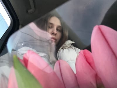 A single female subject is depicted inside a vehicle, viewed through a glass surface. The subject, positioned centrally in the midground, has long, light brown hair and is wearing a light-colored, possibly beige, puffer jacket. Her face is partially visible, with eyes directed towards the camera. In the immediate foreground, a bouquet of several pink tulips with green stems is prominently displayed, partially obscuring the lower and left portions of the female subject. Reflections and distortions are present on the transparent surface, suggesting it is a car window. Visible elements of the vehicle's interior include a dark overhead handle and portions of the ceiling. The scene is located in the city of Slavyansk-na-Kubani, Russia.
