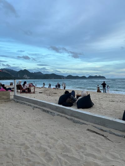 The image depicts a beach scene in Monikeun, Indonesia. The sky is overcast with a mix of dark and light clouds. In the distance, there are several mountainous islands. The beach has a sandy shore leading to the ocean.

Several people are scattered along the beach, some walking by the water's edge and others sitting in chairs or beanbag chairs closer to the foreground. In the foreground, there's a concrete barrier. The overall impression is of a somewhat subdued and casual beach atmosphere.