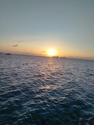 The image depicts a serene seascape at sunset, capturing the tranquil waters of Gulhi Falhu, Maldives. The sun, a bright orb, is setting on the horizon, casting a warm, golden glow across the sky and reflecting on the rippling ocean surface. Several cargo ships are visible in the distance, silhouetted against the vibrant hues of the sunset. A small boat is also present, cruising on the water. The sky transitions from a soft blue at the top to a warm orange and yellow gradient closer to the horizon, with a few wispy clouds adding texture. The foreground is dominated by the deep blue of the ocean, its surface marked by gentle waves.