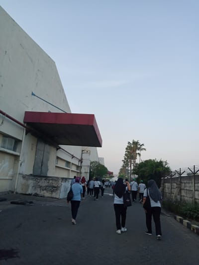 A group of people are walking down a street in Sukomoro, Indonesia. The people are mostly women wearing light blue shirts and dark pants. Some of the women are wearing headscarves. The street is paved with bricks, and there is a tall building on the left and a fence with barbed wire on the right. There are palm trees in the background.