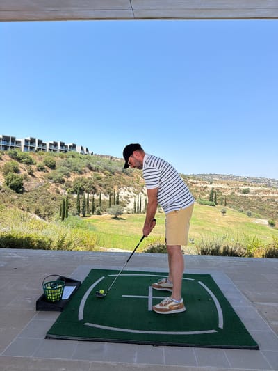 The image captures a man in profile, poised to strike a golf ball on a driving range mat. He is wearing a striped t-shirt, khaki shorts, a black baseball cap, and athletic shoes. Beside him on the mat is a golf ball, and to his left, on a black tray, is a green bucket filled with golf balls. The driving range is set against a backdrop of rolling, arid hills dotted with cypress trees and modern buildings on a hillside. The sky above is a clear, vibrant blue. The scene appears to be in Tsada, Cyprus, given the distinctive landscape and architecture.
