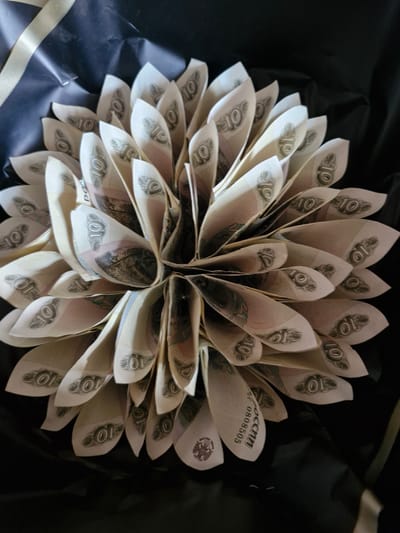 The image displays a bouquet crafted from Russian 10-ruble banknotes, arranged in a circular, petal-like formation. The banknotes are folded and positioned to resemble a flower. The bouquet is presented against a dark, likely black, background, which could be fabric or paper, typical for gift packaging. A light-colored ribbon, possibly gold or cream, is visible in the lower left corner, adding to the gift presentation.
Visible text on the banknotes includes the numeral "10" repeatedly, indicating the denomination. Other visible text includes "России" (Rossii), meaning "Russia," and a serial number "AT 0808505." The banknotes appear to be relatively new, with crisp folds.
The lighting in the image is somewhat dim, creating soft shadows and highlighting the textures of the folded banknotes. The focus is primarily on the bouquet itself, with the background intentionally blurred. The overall impression is that of a creative and monetary gift, possibly for a celebration or special occasion. The image appears to be taken indoors, given the controlled lighting and background. There are no people visible in the media file.