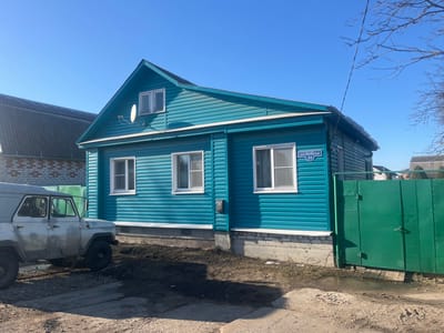 The scene depicts a residential property located at 39 Bebel Street (ул. Бебеля, 39) in Kurbaki, Russia, under a clear blue sky. The primary structure is a single-story house with an attic, featuring horizontal teal-colored siding and white-framed windows. Two large rectangular windows are on the main floor, and two smaller rectangular windows are on the attic level. A small white satellite dish is affixed to the upper facade. The house rests on a white brick or concrete foundation and has a dark-colored roof. A two-panel, solid green metal gate marks the property entrance to the right of the house. On the left side of the frame, a light-colored utility vehicle, resembling a UAZ-469, is parked on a dirt and gravel surface. The vehicle has a soft top and black wheels, and its exterior appears soiled. The ground immediately in front of the house's foundation consists of disturbed earth and muddy areas. Bare tree branches are visible against the sky on the right. No individuals are present, and no active events are occurring.
