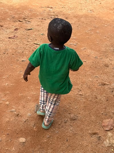 A young child, viewed from behind, walks on a reddish-brown dirt path in Barikin Sabon Gidan Kanat, Nigeria. The child has dark skin and hair and is wearing a green t-shirt with a dark collar and plaid pants featuring pink, blue, and white checkered patterns. The child's feet are clad in light blue slip-on shoes. The ground is covered in small pebbles and dirt, with a few dry leaves scattered around. The lighting suggests it is daytime, with the sun casting soft shadows.