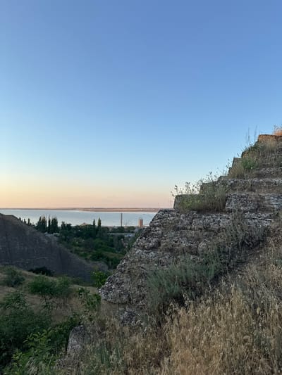 This image, taken vertically, captures a serene twilight scene overlooking a body of water and a distant coastline. The foreground is dominated by a rugged, rocky cliff face, overgrown with dry grass and sparse vegetation. The cliff is textured and appears ancient, suggesting a natural formation or an old structure.
Below the cliff, a dark, stone-built embankment or wall runs along the slope, hinting at historical fortifications or terracing. Beyond this, a vast expanse of water stretches towards the horizon. The water is calm and reflects the soft hues of the setting sun. On the far side of the water, a low-lying coastline is visible, dotted with trees and what appear to be structures, including a tall, slender tower and possibly a bridge in the distance.
The sky transitions from a clear, pale blue at the top of the frame to soft shades of pink and orange near the horizon, characteristic of sunset. The overall mood is peaceful and tranquil, evoking a sense of natural beauty and the passage of time. The lighting indicates it is likely early evening. The image was captured in Mizhlymanske, Ukraine. There is no visible text in the image.