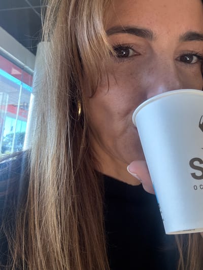 A close-up image depicts a female individual, partially visible from the nose down and extending to the left side of her head, situated in Alcochete, Portugal. She has light brown hair, styled with bangs, and wears a gold-colored hoop earring on her left ear. Her brown eyes are open, with visible dark eyelashes and eyebrows, and freckles are present on her nose and cheeks. She is wearing a dark, possibly black, garment. The individual is holding a white paper cup with her right hand, bringing it to her lips as if drinking. The cup displays a dark logo, with the letter "S" and the letters "O C" visible beneath it. In the background, a large window reveals an exterior scene with a bright sky, reflecting blue and red structural elements, consistent with a commercial building or gas station, and some green foliage. The overall lighting suggests an indoor setting during daylight hours.