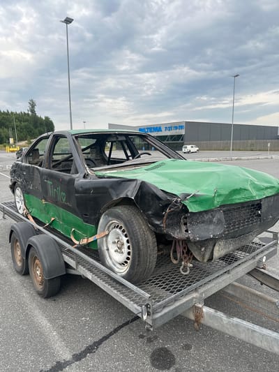 A heavily damaged black and green car is parked on a metal trailer in a paved area. The car has a roll cage visible inside and its hood and front end are severely dented and crumpled. The trailer is attached to a vehicle which is not fully visible. In the background, a large, gray building with "BILITEMA 7-21 (9-19)" written on its facade is visible. There are also streetlights, trees, and a cloudy sky. The car appears to be in a parking lot, likely near the Biltema store. The scene is in Slitu, Norway.