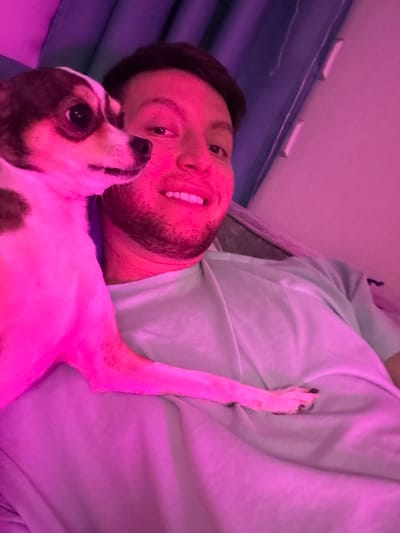 In an indoor setting, possibly a bedroom in Tokaryovo, Russia, a person and a small dog are captured in a selfie-style photograph. The lighting is predominantly pink and purple, casting an unusual hue over the scene. The person, a man with short, dark hair and a beard, is smiling and looking towards the camera. He is wearing a light blue t-shirt. The dog, a small breed with white and brown markings, is positioned close to the man's face, with its paw resting on his chest. The background consists of a blue curtain and a wall, with some light fixtures visible. The overall atmosphere is intimate and relaxed, suggesting a moment of shared affection between the man and his pet.