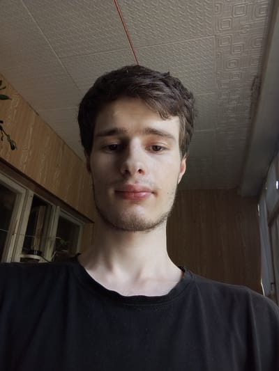 The image is a selfie taken indoors, likely in a residential setting. A young man with brown hair and light stubble is facing the camera, wearing a black t-shirt. The background shows a wood-paneled wall and a ceiling with a tiled pattern. A window with reflections is visible on the left side of the image, and a glimpse of what appears to be a balcony or another window area is on the right. The lighting suggests it is daytime. The location is identified as Nurabad, Uzbekistan.