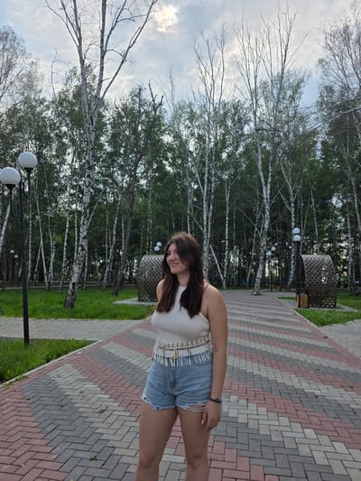 In a park setting in Progress, Russia, a young woman with long brown hair stands on a paved pathway. She is wearing a white sleeveless top and denim shorts. The pathway is laid with interlinking bricks in a pattern of red, grey, and white. To her left, a tall black lamppost with two globe-shaped lights stands near the edge of the pathway. Behind her, a dense grove of birch trees with white trunks and green foliage forms the background. The sky above is overcast with a mix of clouds and some sunlight breaking through. In the distance, several cylindrical, decorative metal structures are visible, one of which appears to be a trash receptacle. The scene is tranquil and appears to be during daytime.