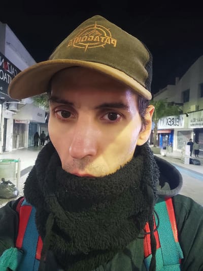A close-up selfie shows a person wearing a brown baseball cap with gold stitching, a dark-colored fuzzy hooded sweatshirt, and what appears to be colorful straps (red, blue, green) underneath. The person's face is the main focus. Outside, the environment shows a street in Lanús Este, Argentina, with buildings and streetlights visible in the background. A sign with mirrored text is discernible. The background is partially blurred due to the shallow depth of field. It is nighttime or low-light conditions.