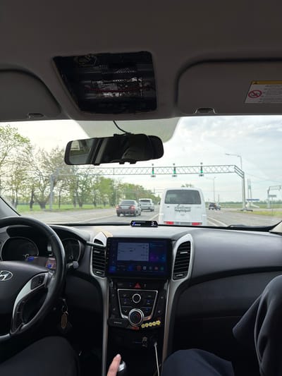 The photograph was taken from inside a car, presumably at a toll gate, with several other vehicles in front. The car's infotainment system shows a map. The dashboard is visible along with the steering wheel with a Hyundai logo. The car is stopped at what appears to be a toll booth in Fedorovskii, Russia. The license plate of the van in front of the car is E910PM 193. There is a Borofone device plugged into the car's console and other objects such as stars and a hanging keychain. There is an opening on the car's roof with wiring visible. The phone displays 17:44 and "Мама, он какой" (Mom, he's so...) in Russian. A finger is pointing toward the BOROFONE device.