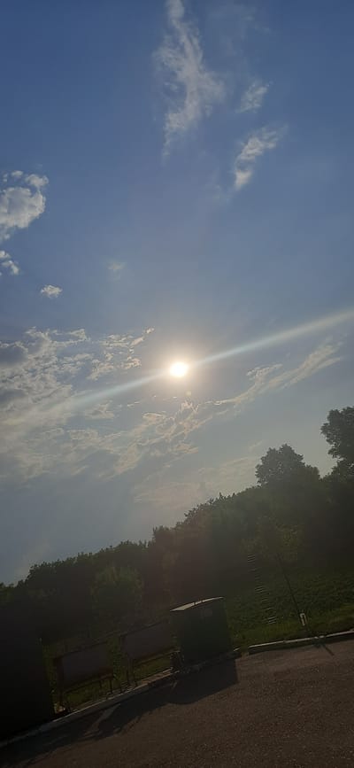 The image depicts a bright, sunlit sky with scattered white clouds. The sun is visible as a strong, glowing orb, casting a beam of light diagonally across the scene. Below the sky, a dense line of green trees forms a backdrop. In the foreground, there is a paved surface, likely a road or path, and a grassy area. A dark, rectangular structure, possibly a trash receptacle or storage bin, is present on the right side of the foreground, along with a bench-like structure. The scene appears to be outdoors, with elements of nature and urban or park infrastructure. The location is identified as Deriivka, Ukraine.