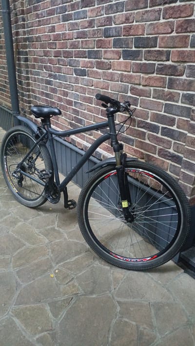 A black bicycle is leaning against a brick wall. The bicycle is a mountain bike with knobby tires, a suspension fork, and disc brakes. It has a black frame, black handlebars, and a black seat. The front wheel of the bicycle is larger than the rear wheel. The bicycle is parked on a cobblestone patio. The brick wall is made of red and brown bricks. The scene is outdoors, and it appears to be daytime. There is no visible text in the image. The location is Nikolino, Russia.