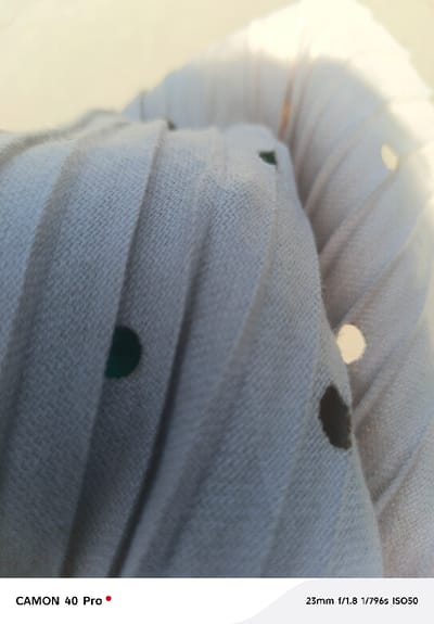 The image depicts a close-up, out-of-focus view of a light-colored, possibly off-white or light grey, pleated fabric. The fabric exhibits a visible woven texture and is adorned with multiple circular markings, appearing as polka dots, in varying shades of dark green, dark brown/black, and one bright white. The pleats run vertically across the frame, creating distinct lines and shadows. A strong, bright light source, likely natural light, originates from the upper right, causing parts of the fabric to appear overexposed and casting soft highlights. The background is completely blurred due to a shallow depth of field. No individuals are present. No specific actions or interactions are depicted. The environment is limited to the fabric and ambient light conditions. The image is stated to be from Nakuru East ward, Nakuru City, Kenya. Camera metadata "CAMON 40 Pro" and "23mm f/1.8 1/796s ISO50" are visible at the bottom.