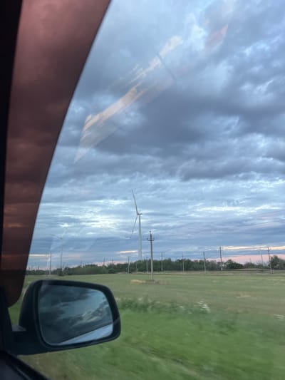 A side view mirror reflects a cloudy sky with subtle pink hues, suggesting either dawn or dusk. The mirror itself is attached to the frame of a vehicle, with a dark, possibly red-tinted interior visible along the left edge. Beyond the mirror, the landscape unfolds with a field of green grass extending towards a line of trees and power lines in the distance. Several wind turbines are scattered across the horizon, some with their blades visible, indicating a wind farm. The overall impression is one of a rural or semi-rural environment, possibly on the outskirts of Zimovniki, Russia, given the presence of wind turbines and open fields. The lighting suggests an overcast day or twilight hours.