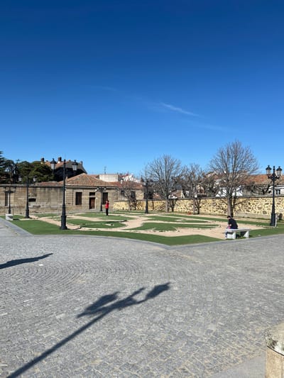 The image displays a sunny public square in Piedrahíta, Spain. The foreground is paved with grey cobblestones arranged in a fan pattern, marked by long shadows from streetlights and an unidentifiable source. The midground features a rectangular garden containing patterned sections of green grass separated by sandy paths. Black, ornate street lamps with two lanterns are situated around the garden perimeter and on the surrounding pavement. One person, dressed in a red top and dark pants, stands in the garden's left section, facing right. Another person, in dark clothing, is seated on a stone bench on the right side of the garden, facing left. Behind the garden, a low stone wall precedes several buildings with terracotta tiled roofs and light-colored facades. Bare deciduous trees and some evergreen foliage are visible in the background against a clear blue sky with minimal cloud cover.