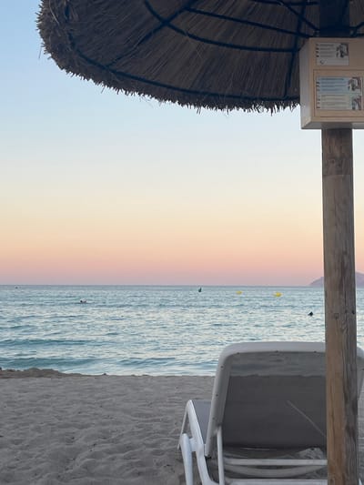 This image depicts a serene beach scene at Platja D'alcúdia, Spain. The focus is on the ocean and the sky, with elements of beach furniture and a thatched umbrella framing the view.
In the foreground, a portion of a large thatched umbrella is visible on the left, suggesting a shaded area on the beach. To the right, a white beach chair is partially shown, indicating relaxation and leisure. A wooden beam crosses the upper part of the frame, likely part of a beach structure or pergola.
The ocean takes up the right side of the image, its surface rippled with gentle waves. A few small, colorful buoys are scattered in the water, and a couple of distant figures can be seen wading or swimming. The sand of the beach is visible at the very bottom right.
The sky transitions from a pale, almost white hue at the horizon to soft pastel colors of pink and orange higher up, suggesting either sunrise or sunset. This gradient of color in the sky creates a peaceful and tranquil atmosphere.
There are two small signs attached to a light-colored rectangular box, which is mounted on the wooden beam. The text on these signs is not clearly legible but appears to be informational, possibly displaying rules or information about the beach facilities.
The overall impression is one of a calm and beautiful day at the beach, with the soft colors of the sky and the gentle movement of the water contributing to a feeling of peace.