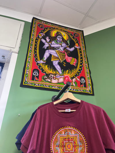 An indoor scene, possibly a shop or decorated room in Sondrio, Italy, displays cultural and spiritual-themed items. The dominant feature is a vibrant, rectangular tapestry or poster hanging on a light green wall. It depicts the Hindu deity Shiva Nataraja in a dynamic, dancing pose, rendered in shades of purple and lavender with multiple arms, flowing hair, and traditional attire, surrounded by a fiery golden halo. Below the main figure, a smaller, inverted figure, possibly Apasmara, is shown. The tapestry features a red border with intricate patterns, including black tridents (Trishula) and golden Om symbols.
To the right of the tapestry, a maroon t-shirt hangs on a wooden hanger, suspended from a black metal pipe rack affixed to the green wall. The t-shirt prominently features a large, colorful, circular mandala design on its front, composed of intricate geometric patterns in shades of red, orange, yellow, and blue, with an Om symbol at its center. A small white label or tag is visible on the t-shirt's collar, though its text is unreadable. Partially visible beneath the maroon shirt is a dark blue t-shirt. The green wall continues to the right, and to the left, a white wall or ceiling section with subtle paneling is visible, forming a corner with the green wall. The lighting appears even, suggesting indoor artificial light.