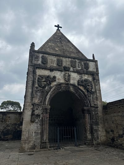 This image shows the exterior of a stone structure, likely a chapel or shrine, with a prominent arched entrance. The structure is made of weathered stone blocks and features intricate carvings. Above the arch, there are depictions of angels holding candelabras and shields with coats of arms. A cross is visible at the apex of the building's triangular gable. The area in front of the entrance is paved with stone, and a dark metal gate is positioned within the archway. To the left of the structure, a stone wall is visible, and further in the background, a single tree stands against a cloudy sky. The overall impression is of an old, historic religious site. This structure is located in Huejotzingo, Mexico, and is part of the Franciscan monastery complex.