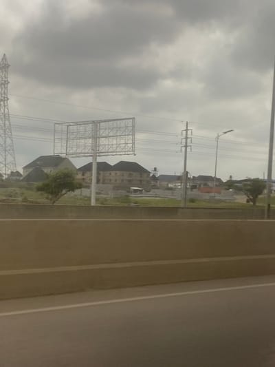 The image, oriented horizontally, depicts an outdoor scene observed from a perspective that suggests looking out from a vehicle or behind a fixed structure.
In the right foreground, a broad, light-colored surface with prominent vertical lines runs vertically through the frame, possibly representing a road, a highway barrier, or a part of the immediate viewing enclosure.
The left and central portions of the midground reveal an urban or suburban landscape. Numerous utility poles with extensive power lines crisscross the area, one featuring a streetlight. A large, skeletal metal billboard frame stands conspicuously in the mid-left, currently empty without any advertisements. Below and beyond these structures, several multi-story buildings with dark, sloped roofs are visible, some appearing residential or commercial. Sparse green trees and patchy vegetation dot the landscape between the buildings and beneath the power lines. The ground appears to be a mix of undeveloped earth and grass.
The background is dominated by an extensive, uniformly light grey and heavily overcast sky, indicating a gloomy or cloudy daytime.
No specific activities or people are discernible in the scene. The weather is characterized by heavy cloud cover. There is no visible text. The environment suggests an area undergoing development or an established infrastructure-heavy zone, consistent with a location like Gosa, Nigeria.